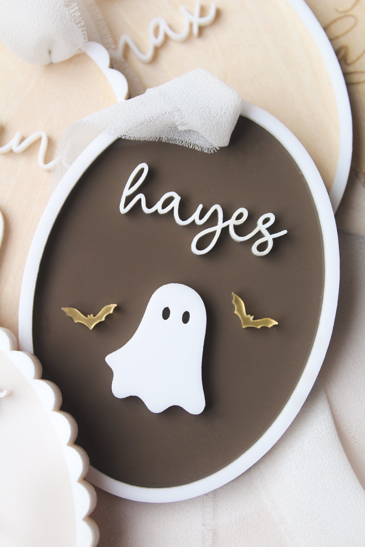 A close-up of a round decorative plaque or ornament with a dark brown center, a white border, a white ghost cutout, two small gold bat shapes, and the word 'hayes' in white script near the top.